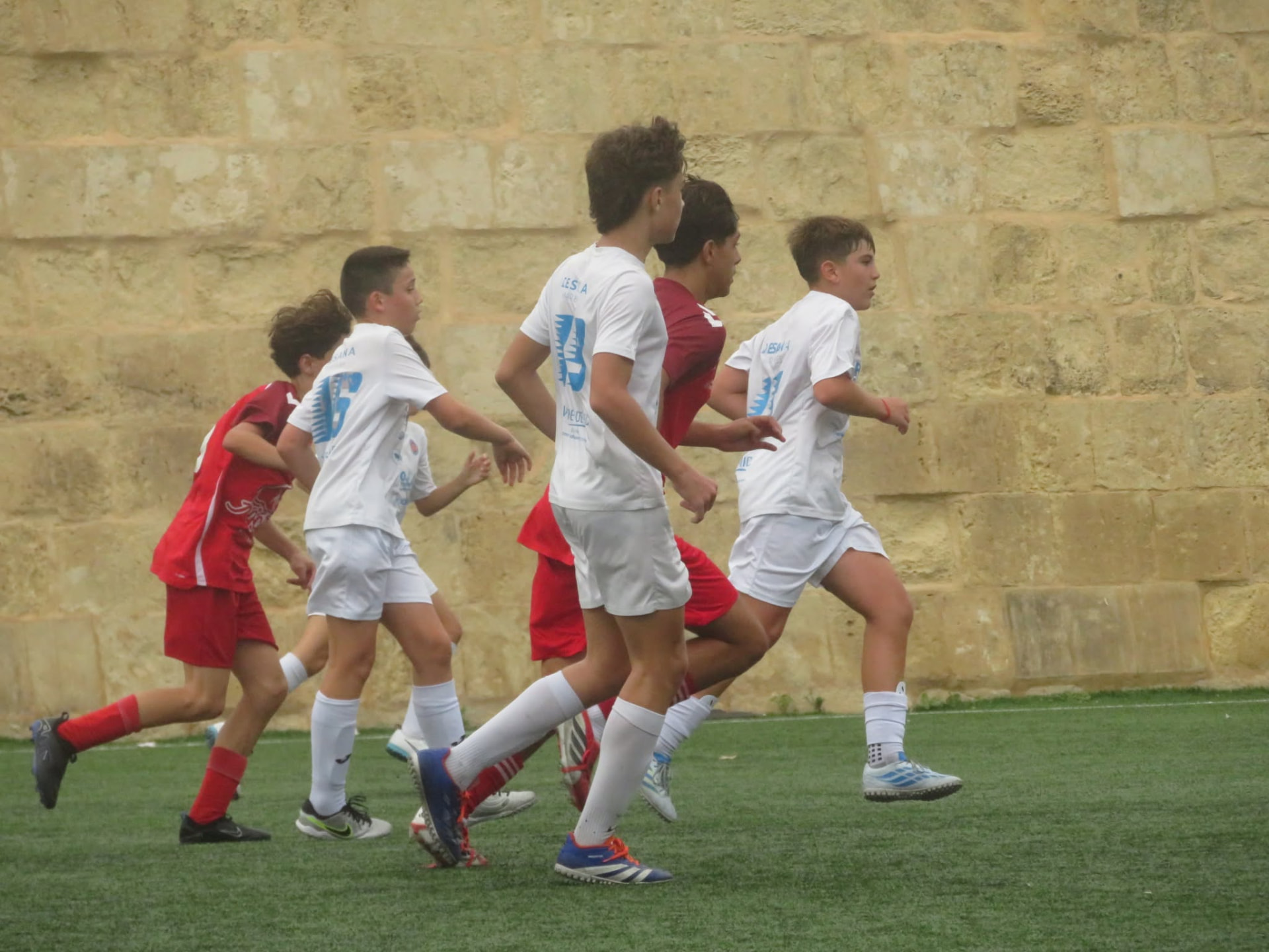 Grupp ta' plejers żgħażagħ bl-abjad u l-aħmar jiġru flimkien quddiem ħajt kbir tal-ġebla Maltija.