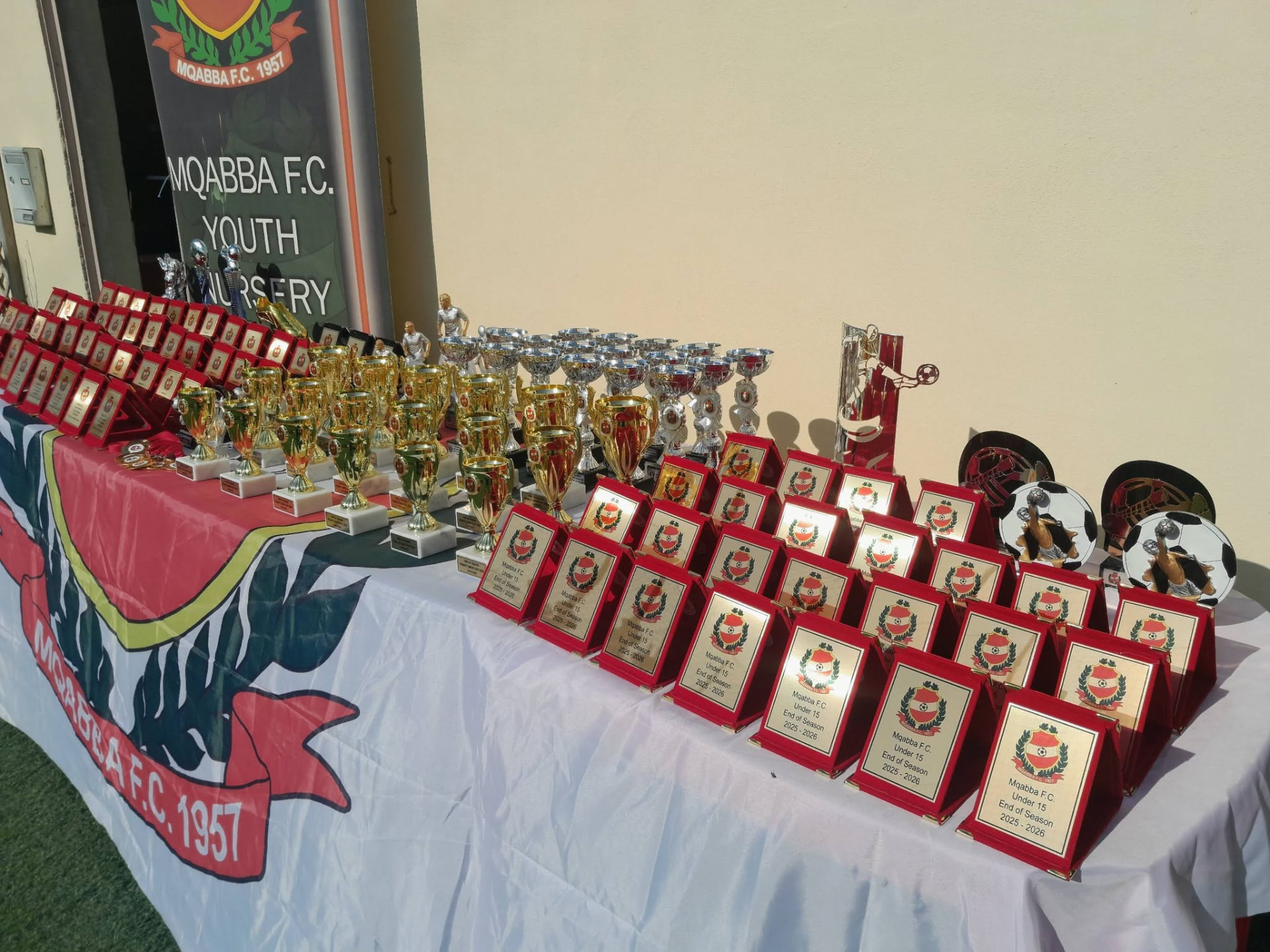 Mejda mimlija trofej u plakek ta' Mqabba F.C. Under 15 għall-għeluq tal-istaġun 2025–2026, quddiem banner tal-Youth Nursery.